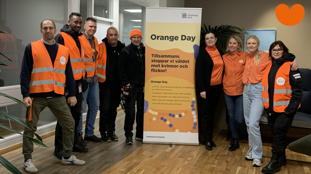 9 personer i orangea västar tittar mot kameran och ser innerliga ut. I mitten en Orange Day-vepa med liten text. Ett orange hjärta längst upp till höger. De står inne i entrén till distriktskontoret