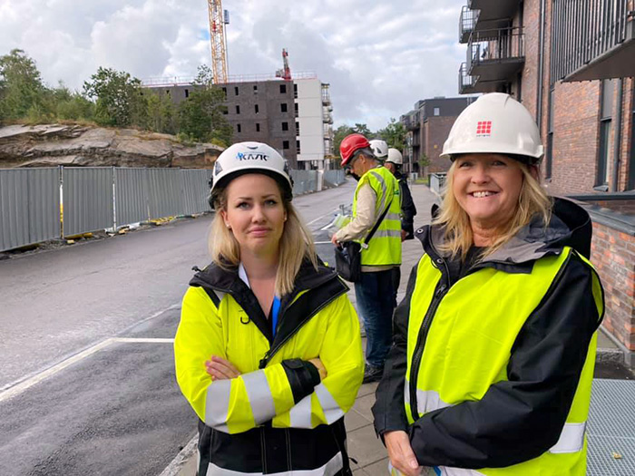 Två kvinnor med bygghjälmar och byggvästar. I bakgrunden hus och människor som också inspekterar bygget.,